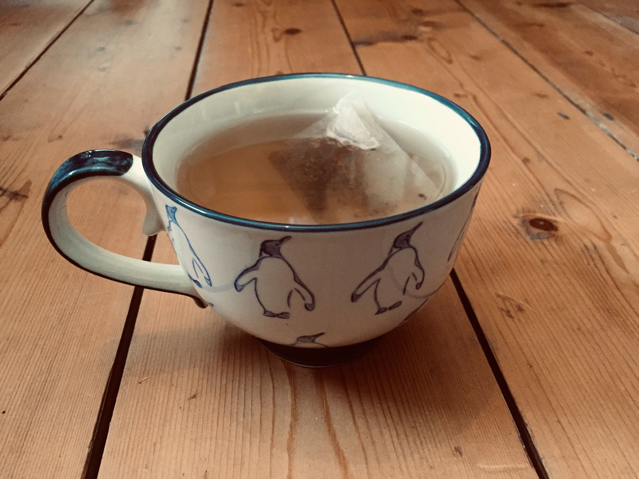 A cup decorated with penguins, containing a teabag in hot water, on wooden planks